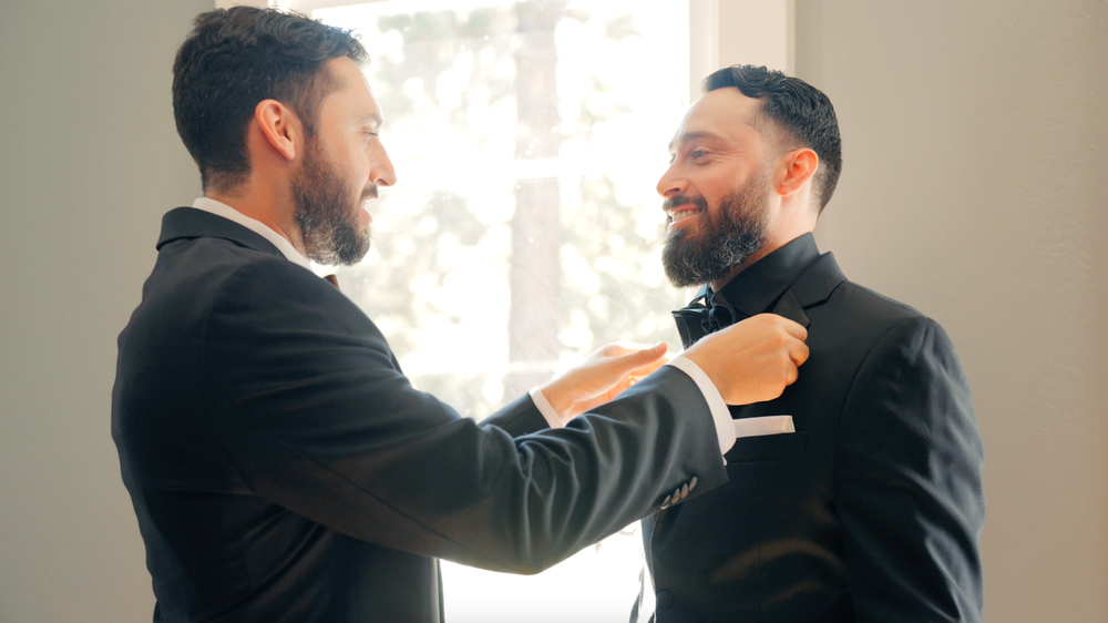 Two people in black suits stand close together adjusting each other's formal attire before a celebration.