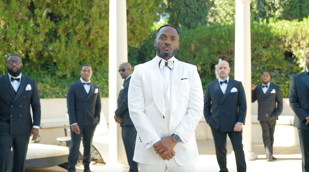 A wedding party in formal attire poses outdoors with the groom in white.