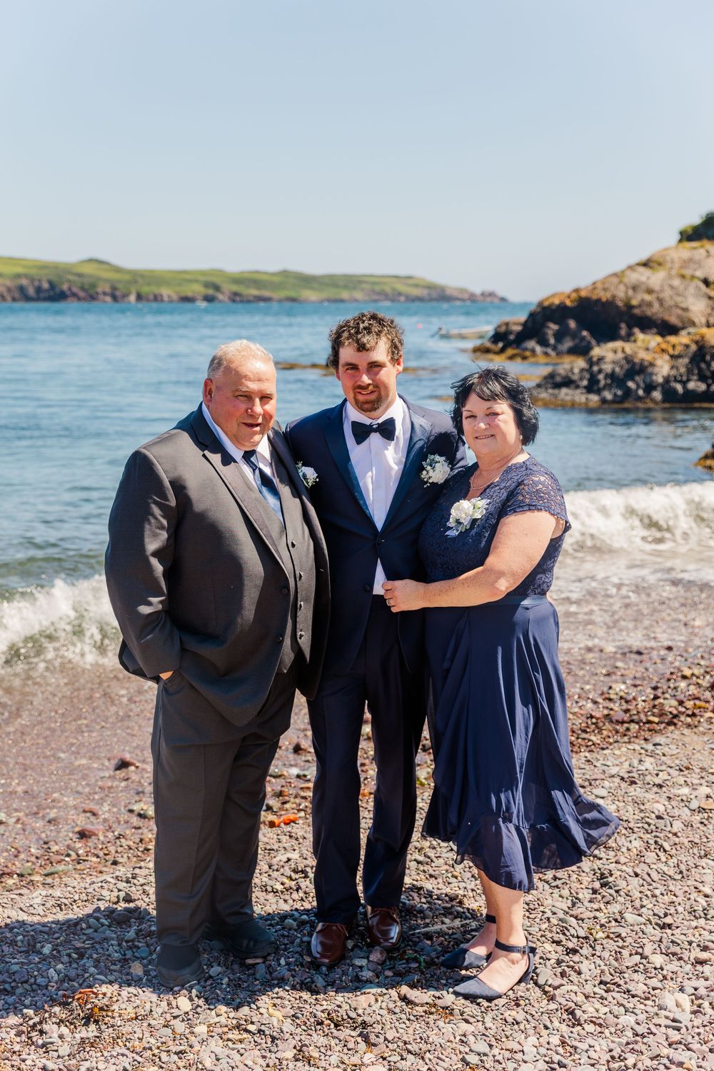 Wedding party members stand together on a rocky beach with coastal scenery behind them.