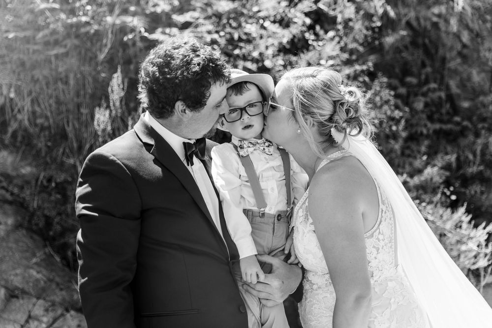 Black and white outdoor wedding photo showing couple with their young child standing between them in nature setting.