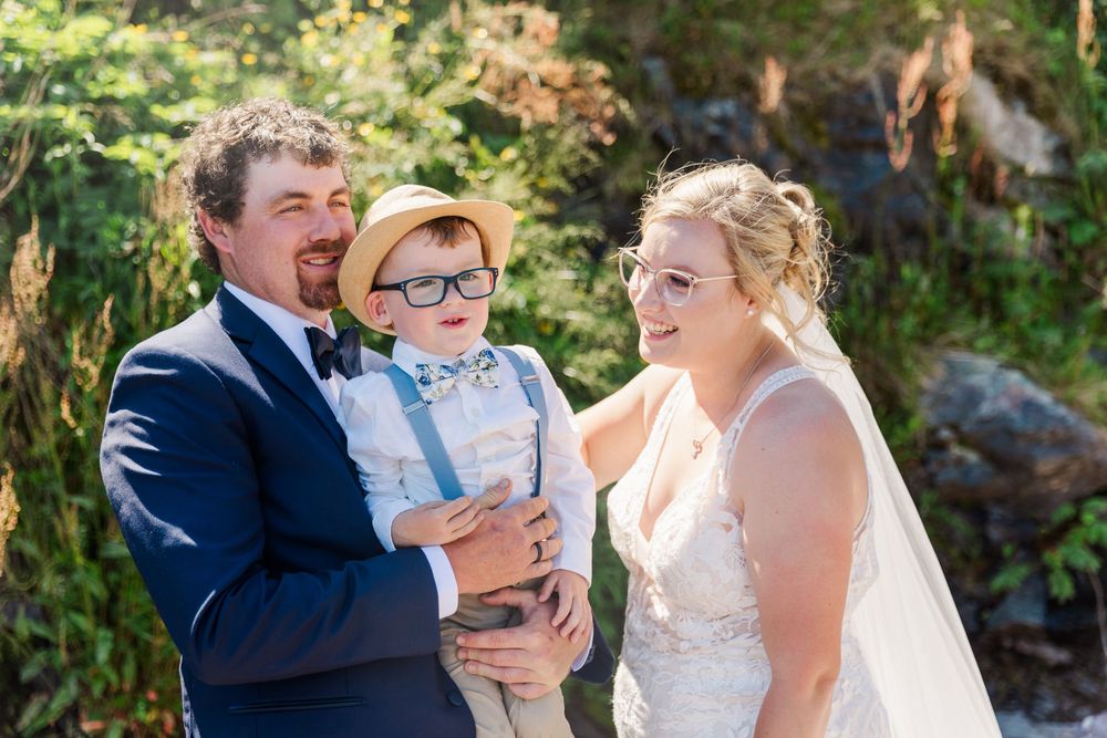 A joyful family moment at a wedding shows the bride and groom with a small child wearing glasses and a bowtie outdoors.
