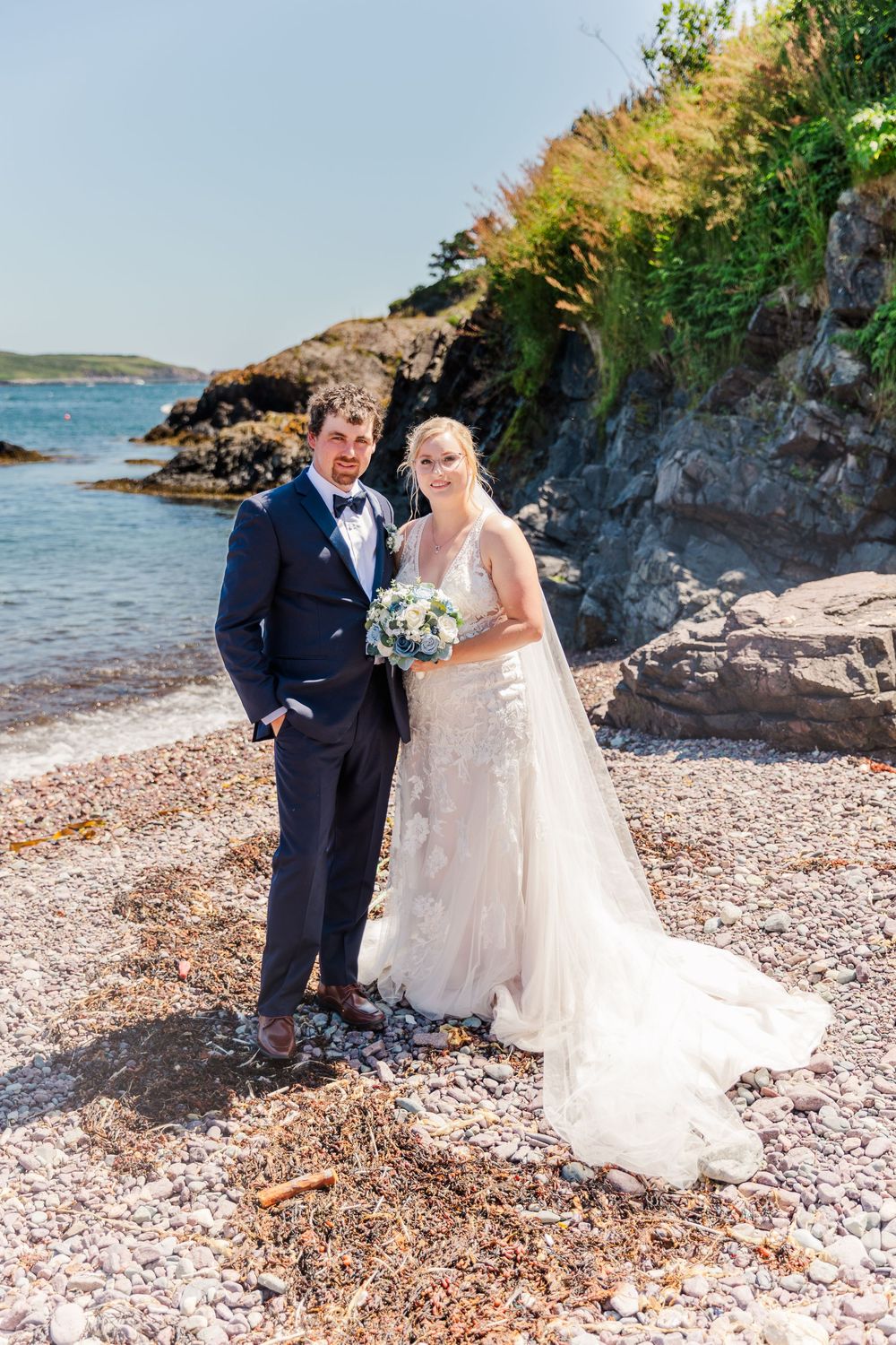 A wedding couple poses on a rocky beach with ocean and cliffs in the background on a sunny day.