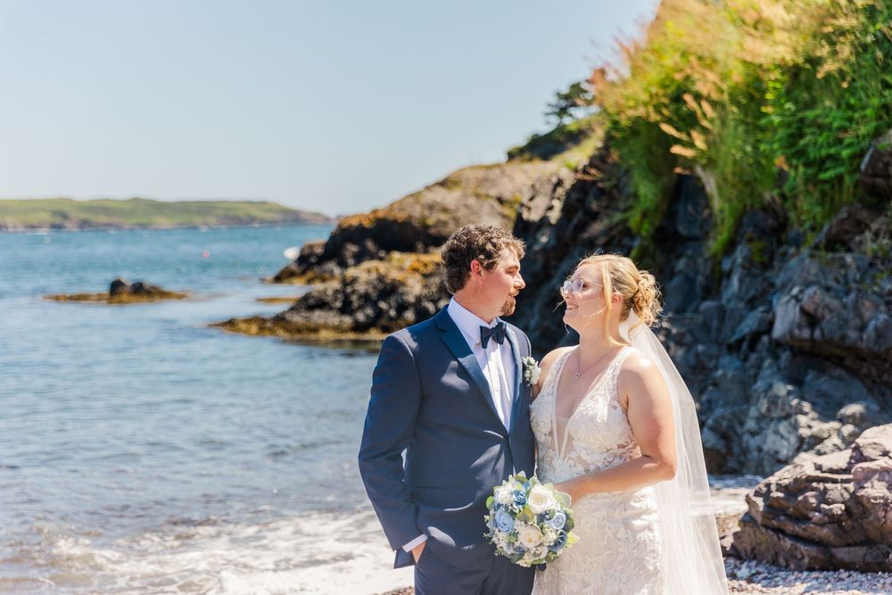 A romantic coastal wedding portrait with a couple posing against rocky cliffs and blue ocean waters on a sunny day.