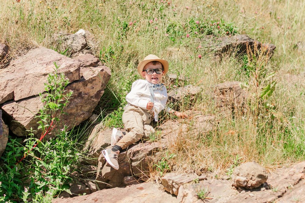 Someone in a straw hat and light clothing sits on rocky terrain surrounded by dry grass and boulders in a natural setting.