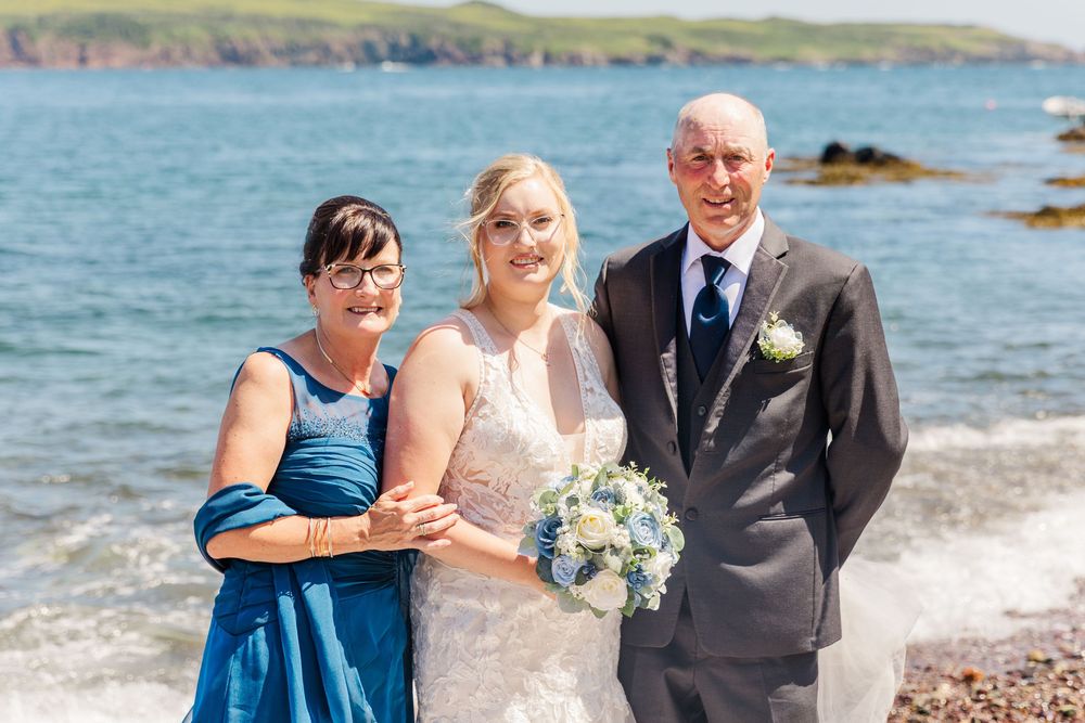 Three people in formal attire pose together by the ocean shore on a sunny day with blue water and cliffs in background.