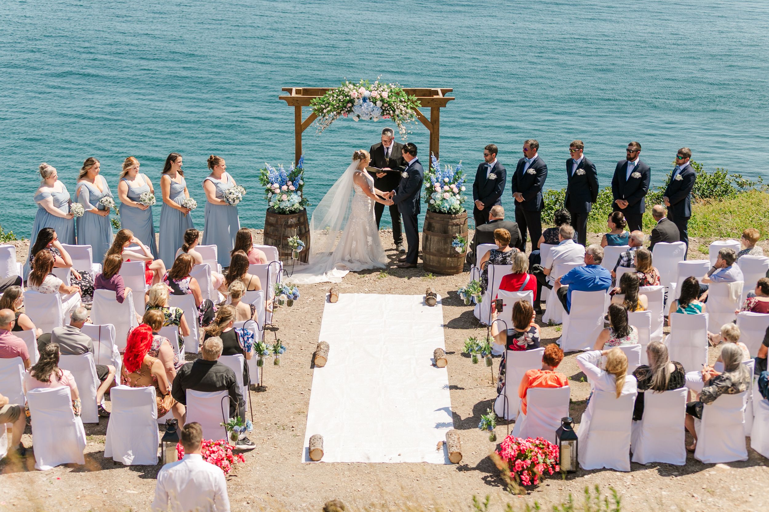 A wedding ceremony takes place on a cliffside overlooking turquoise waters with guests seated in white chairs.
