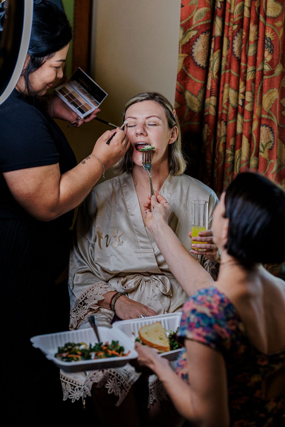 Wedding preparations showing stylists applying makeup in a room with floral curtains.