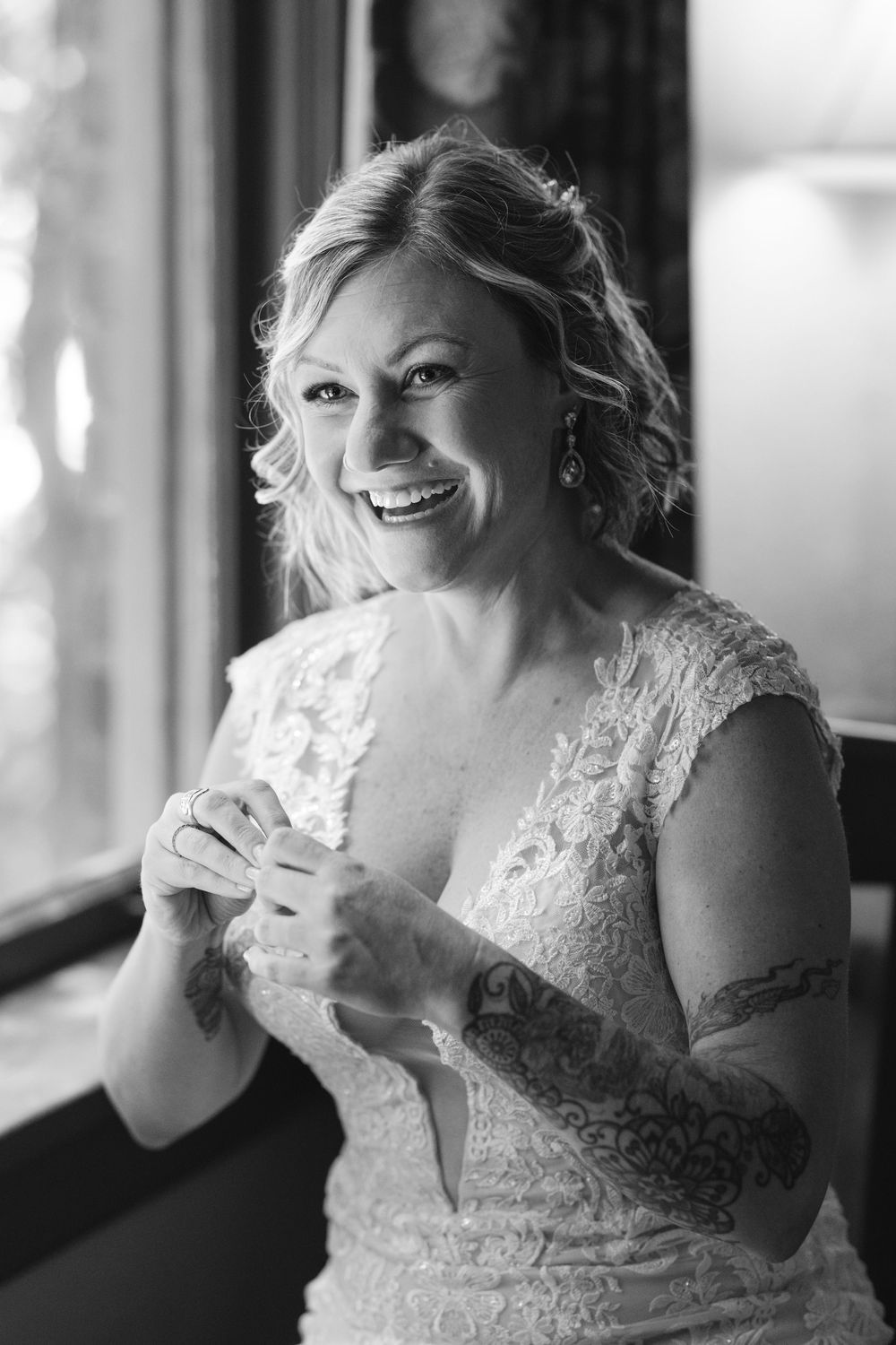 Black and white portrait of a bride in a lace wedding dress laughing joyfully near a window.