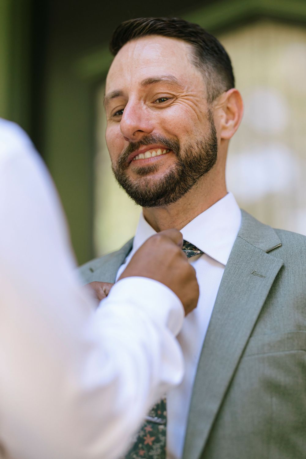 Bearded groomsman in grey suit jacket smiles warmly during outdoor wedding ceremony.