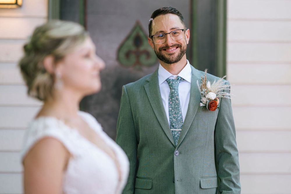 A wedding couple in elegant attire poses outdoors, with a sage green suit and white dress creating a classic look.