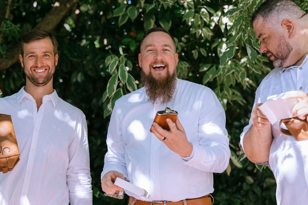 Wedding party in white shirts laughing and sharing drinks outdoors in dappled sunlight.