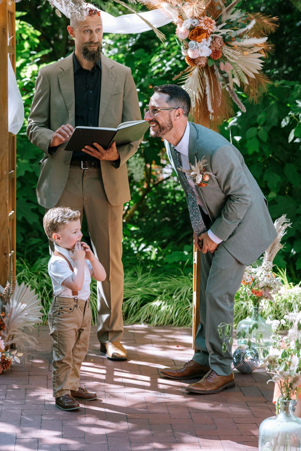 A young child playfully interacts with someone in a suit during an outdoor wedding ceremony under a wooden pergola.