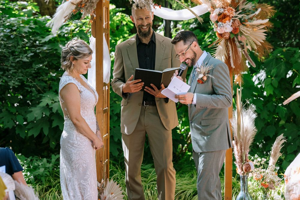 Wedding ceremony taking place under a wooden arch decorated with pink and peach flowers in an outdoor garden setting.