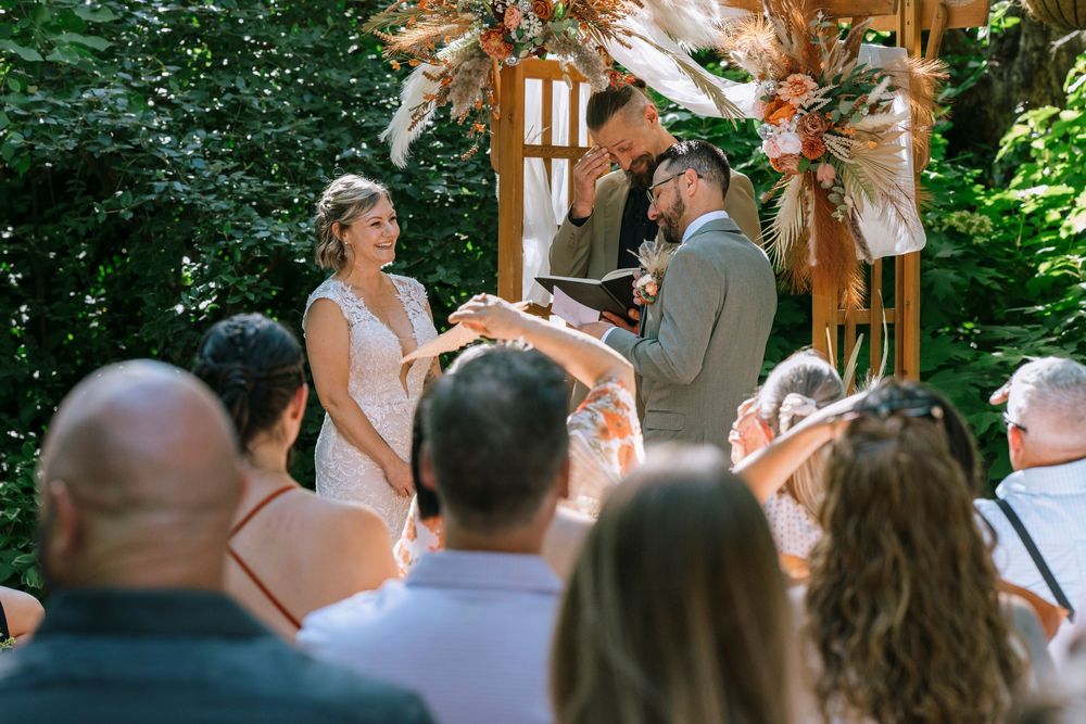 People gather around a decorated wooden arch during an outdoor wedding ceremony surrounded by lush greenery.