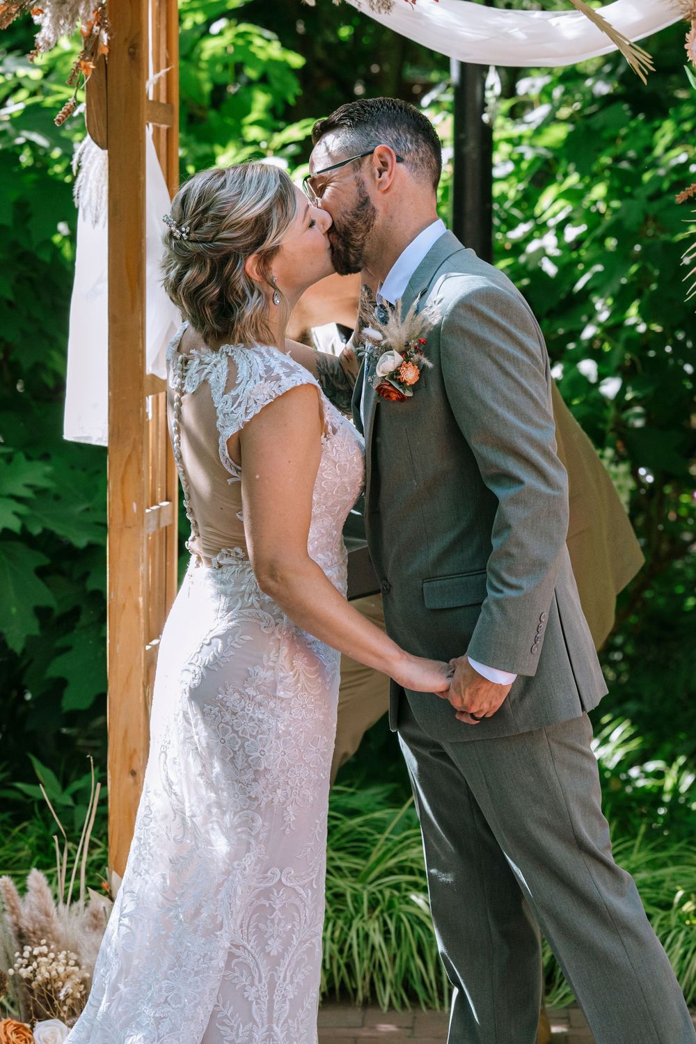 A romantic first kiss between newlyweds under a wooden gazebo surrounded by lush greenery on their wedding day.