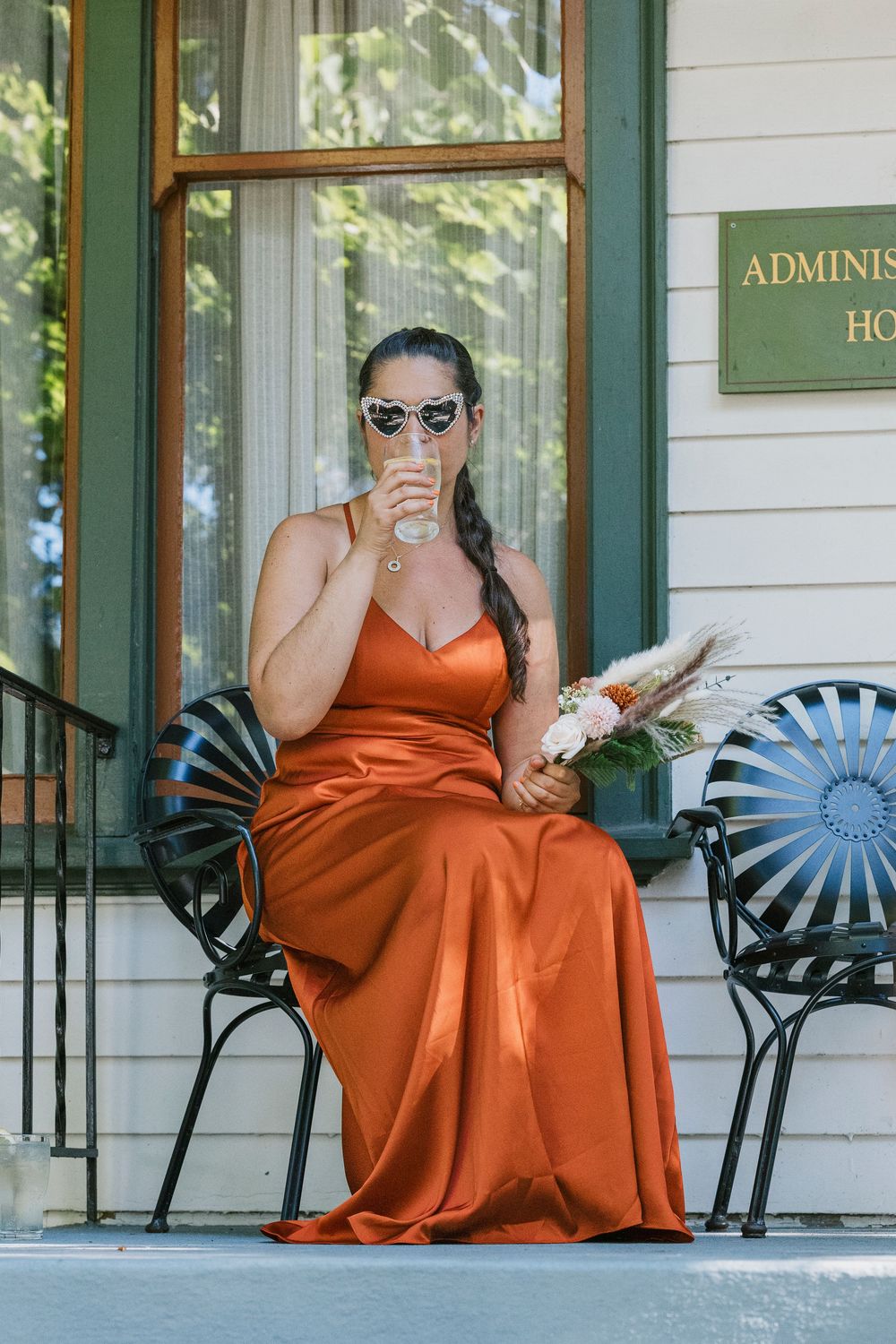 A person wearing an orange dress relaxes in a black metal chair outside a white building with green trim.