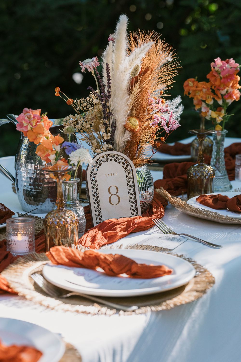 Elegant table setting with orange napkins, pampas grass centerpieces, and rustic place settings on a white tablecloth.