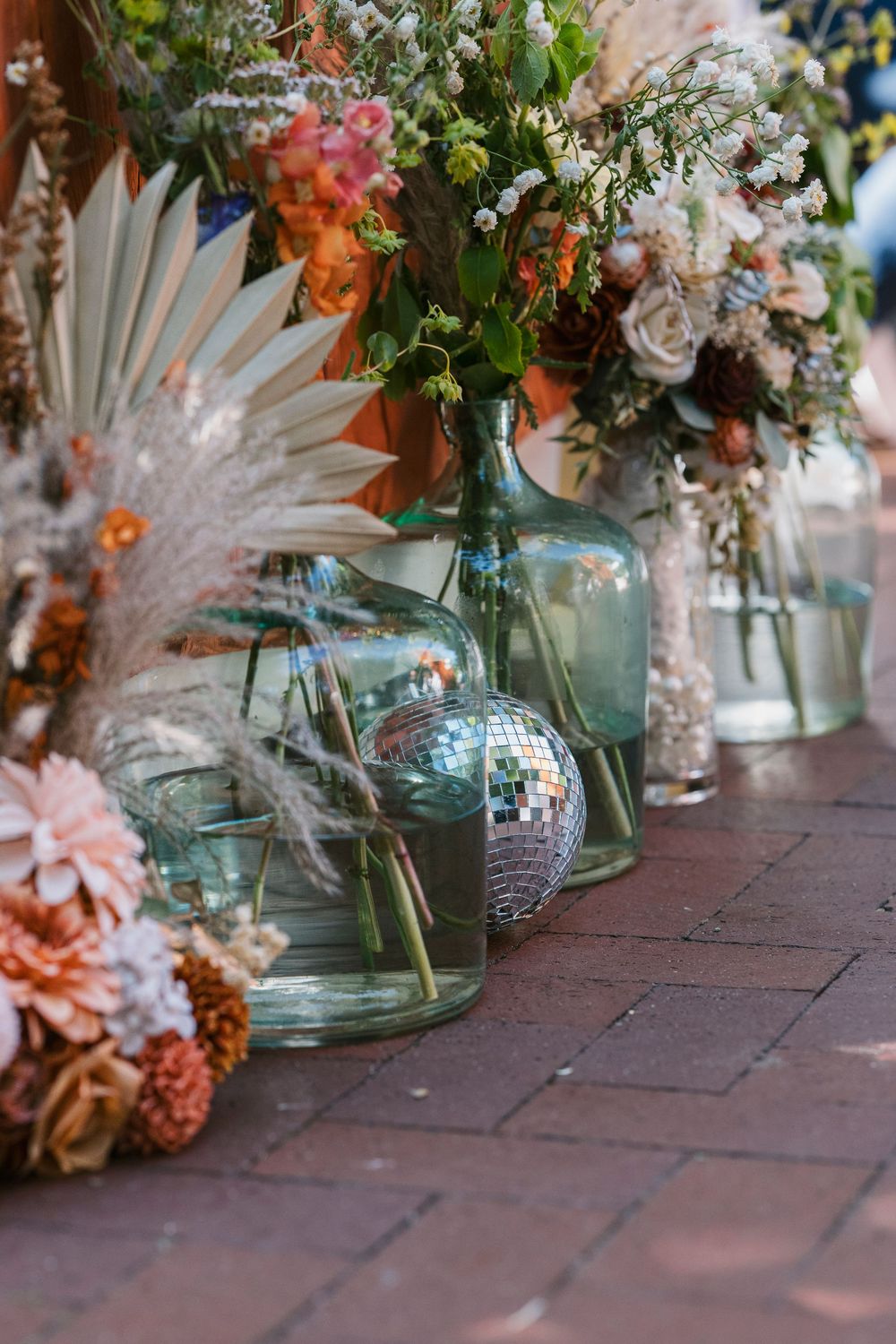 Row of glass vases with colorful floral arrangements and white feathers sit on brick surface.