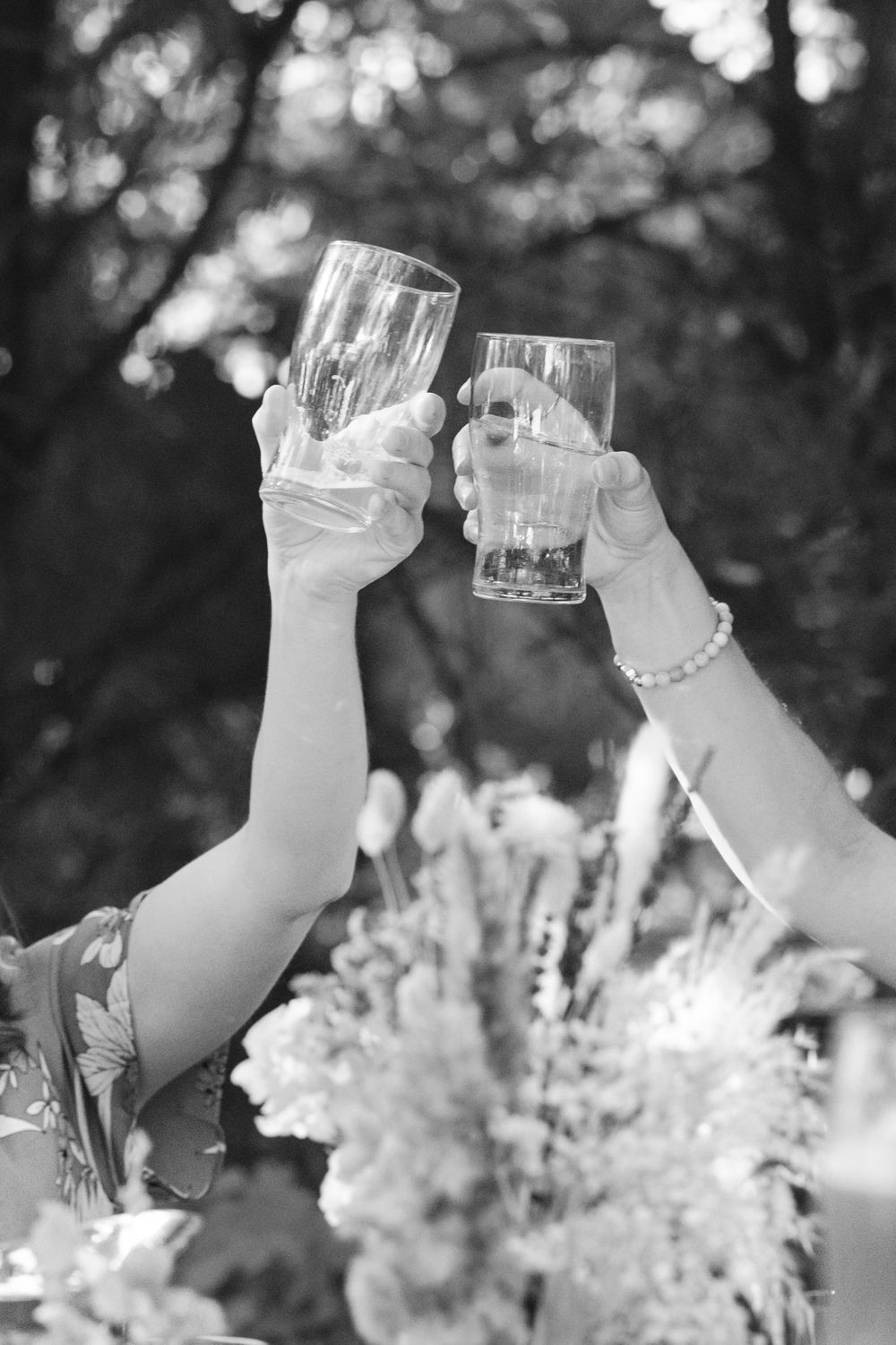 Two hands raise champagne glasses in a celebratory toast against a blurred outdoor background in black and white.