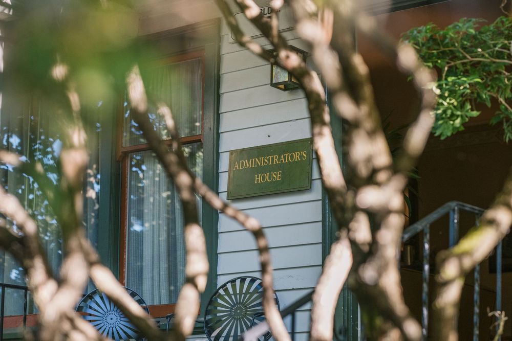 A vintage-style administration house entrance with scrollwork details viewed through blurred tree branches in the foreground.