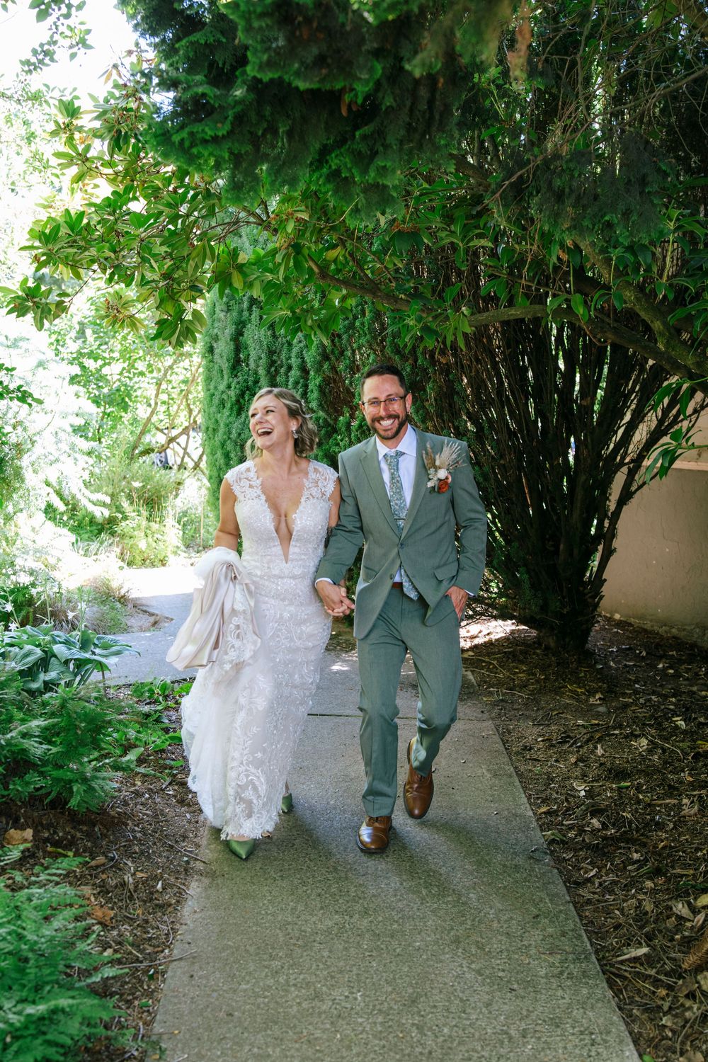 A couple in wedding attire walk hand in hand along a garden path under lush greenery and hanging vines.