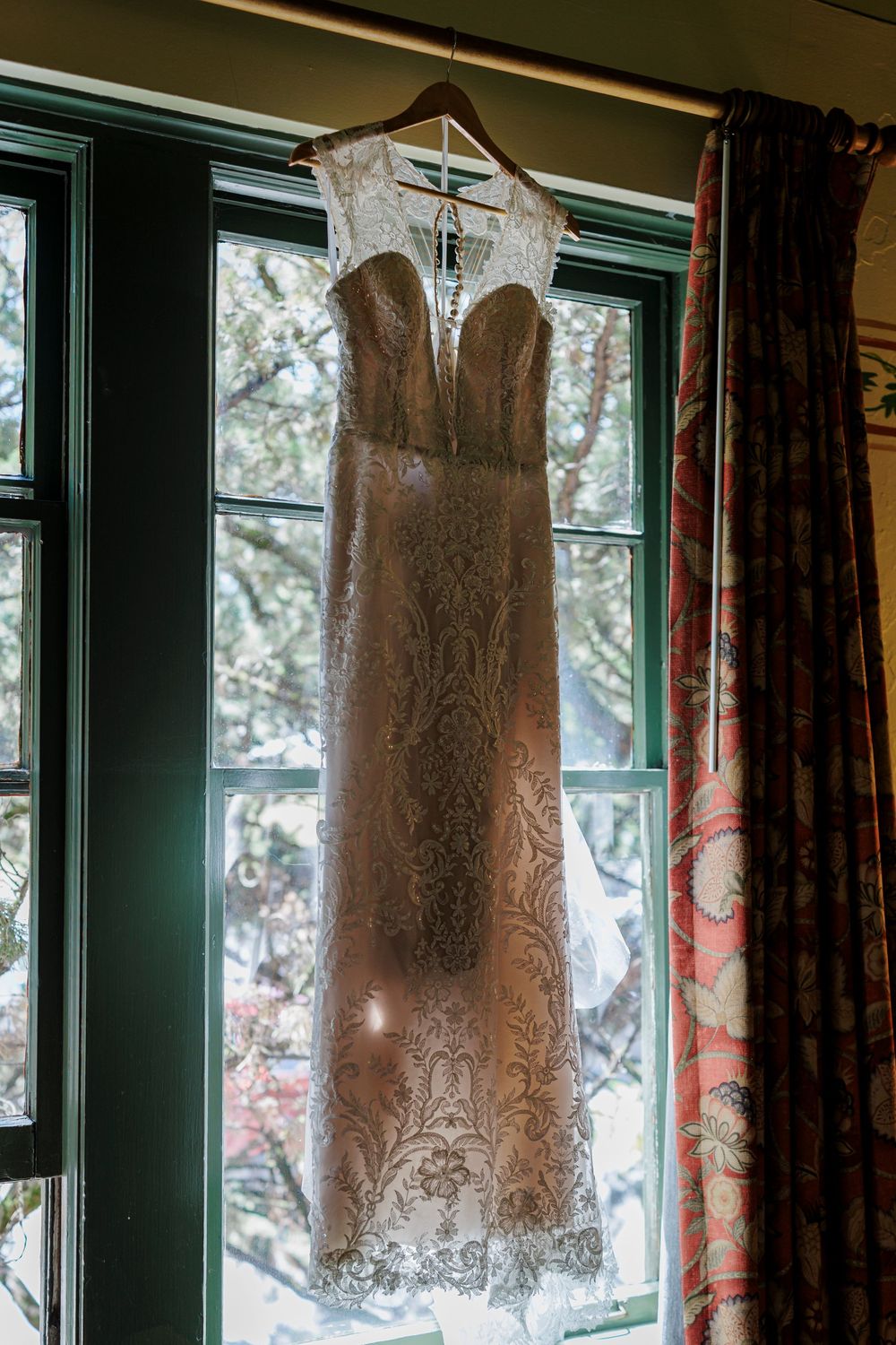 Elegant lace wedding dress hanging in front of a window with green trim and decorative curtains.