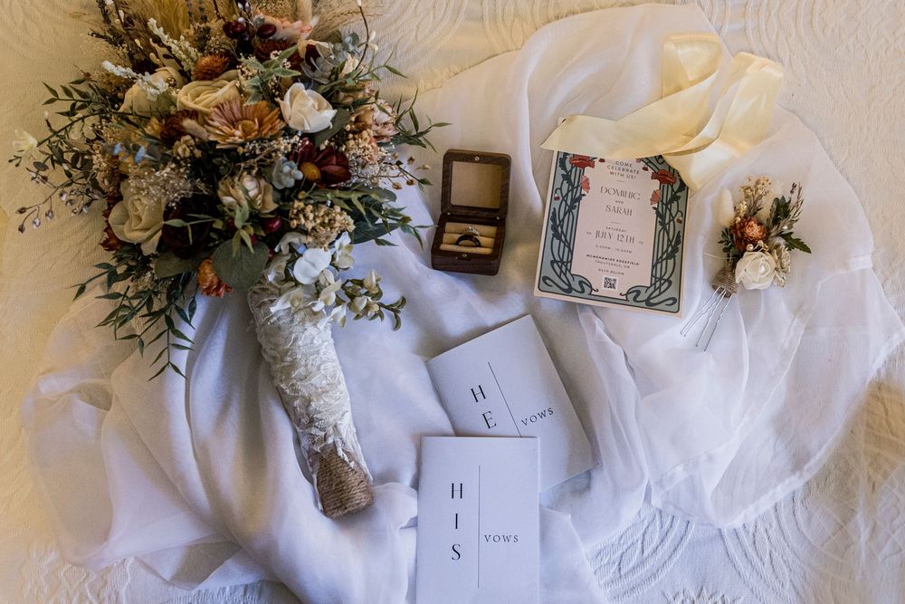Wedding details flat lay with bouquet, rings, and stationery on white fabric.