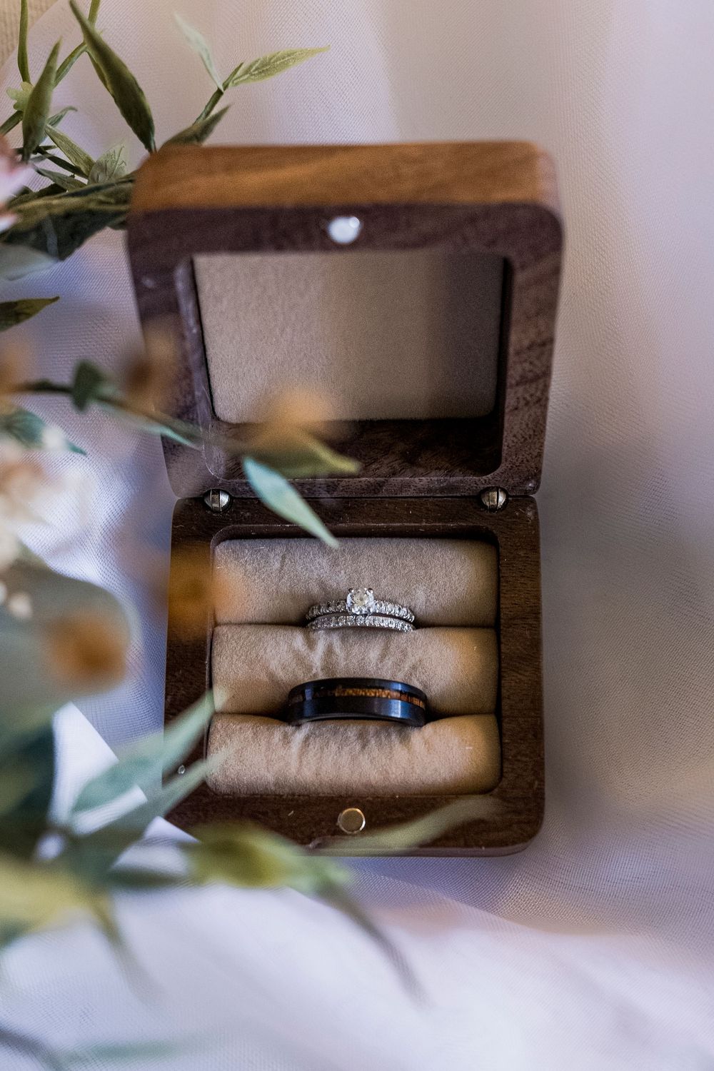 Wedding rings nestled in a wooden box with greenery in soft focus.