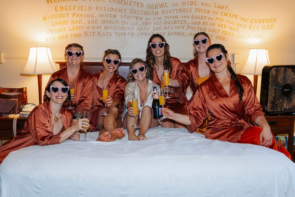 Wedding party wearing matching orange robes and sunglasses posed together on a bed.