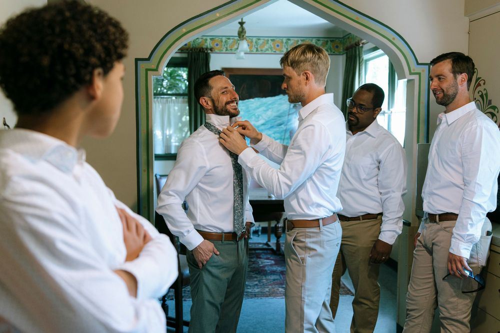 Groomsmen helping adjust each other's ties while getting ready in a room with an arched doorway.