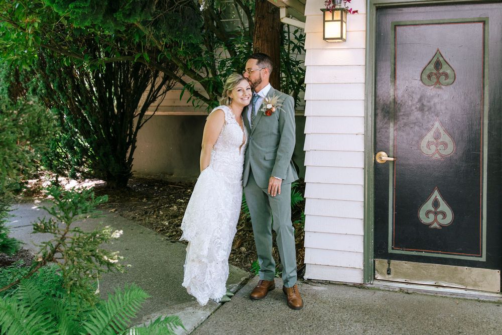 Couple in wedding attire poses against white building with spade-decorated black door.