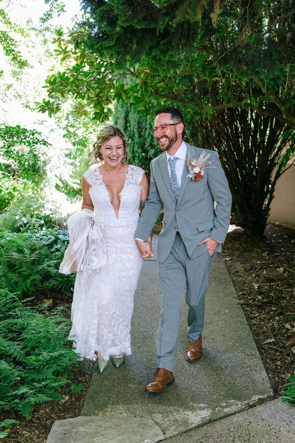 A couple walking hand in hand on a garden path, wearing wedding attire amidst green foliage.