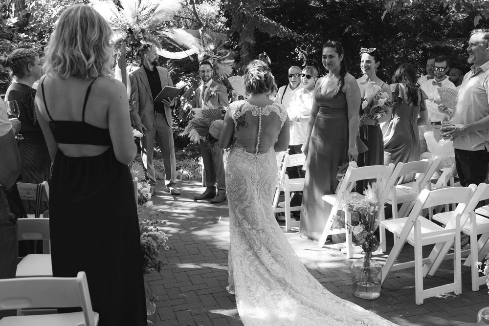 Wedding guests seated in white chairs at an outdoor ceremony beneath shady trees.