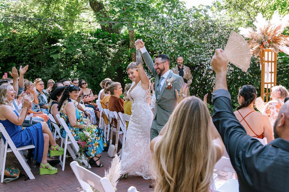 Outdoor wedding celebration with guests cheering as the couple walks down the aisle under shaded trees.
