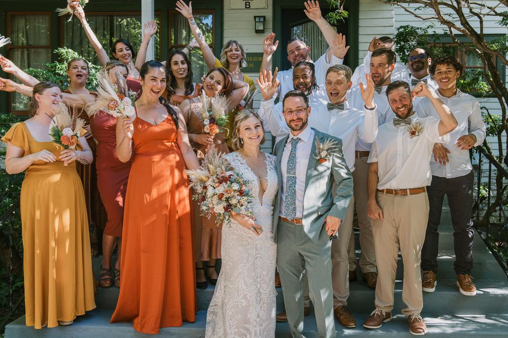 Wedding party dressed in tan suits and orange dresses celebrate outdoors on a sunny day.