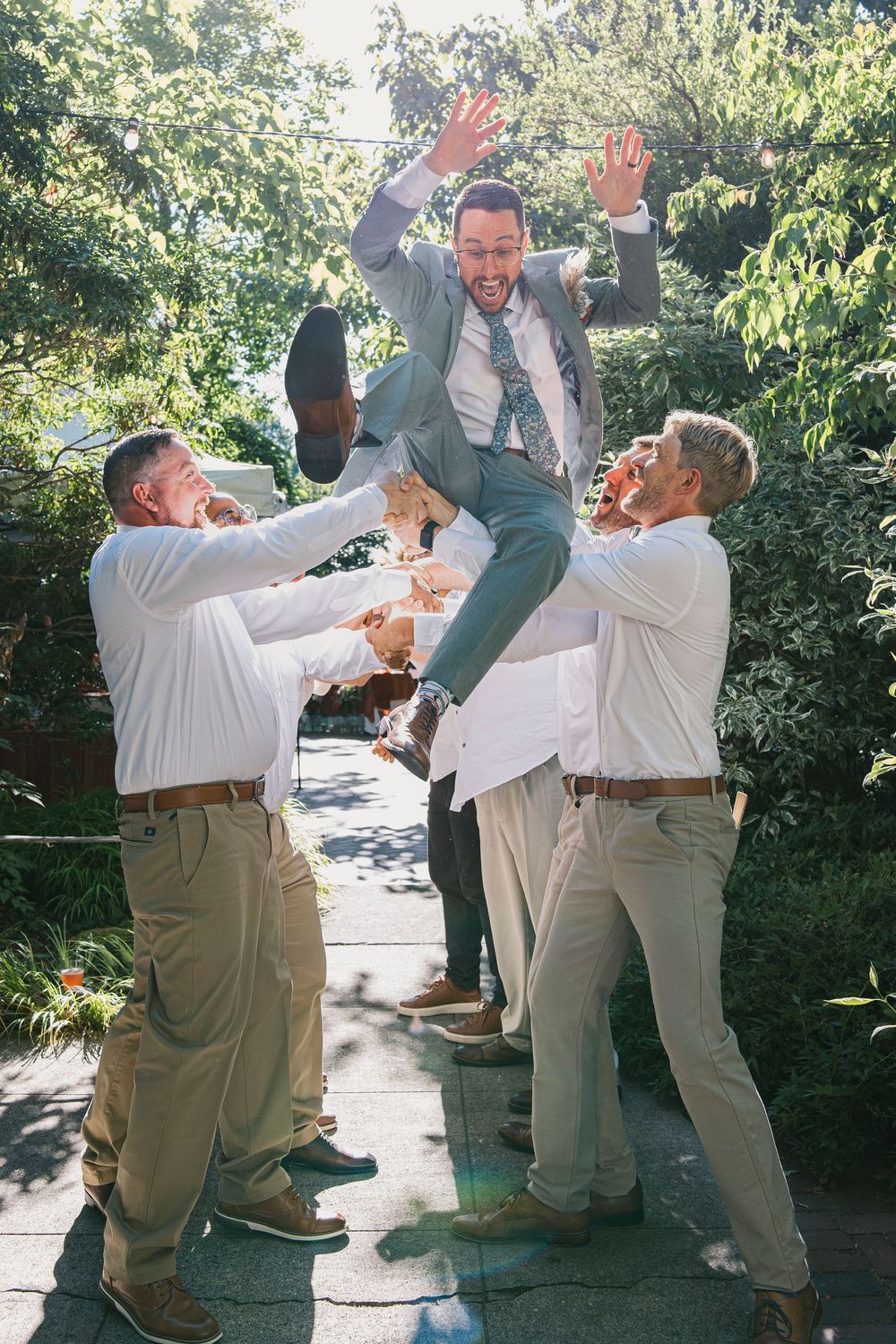 Groomsmen in khaki suits perform playful jumping poses for photos in a garden setting.
