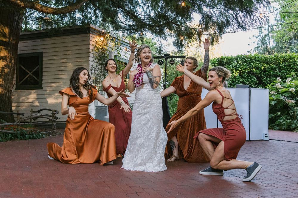 Wedding party poses playfully outdoors in rustic setting with bridesmaids wearing burnt orange dresses and white bridal gown.