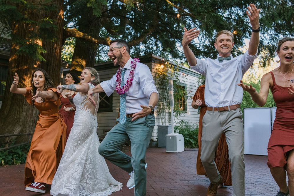 A group dances joyfully at an outdoor celebration under trees at sunset, showing lively movement and celebration.