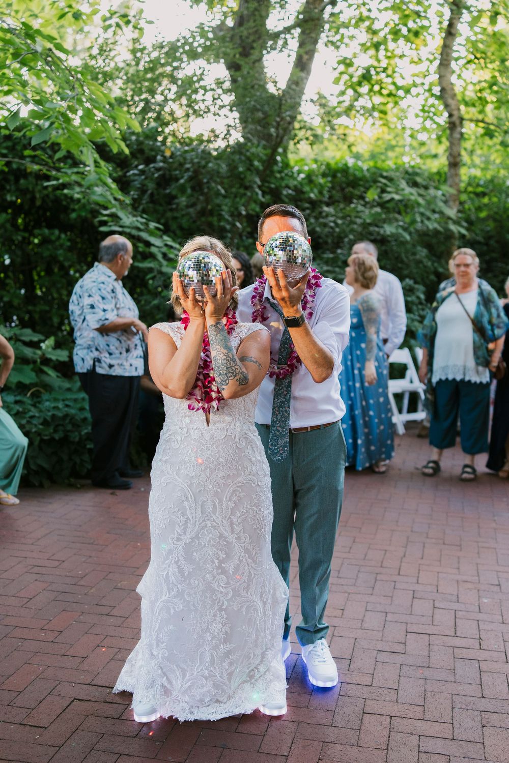 A couple shares their first drink at an outdoor garden wedding reception on a brick patio with guests in the background.