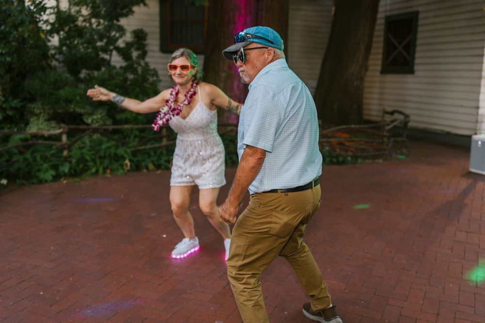 Two people dance together on a brick patio outside a house, one wearing khakis and the other in white shorts.