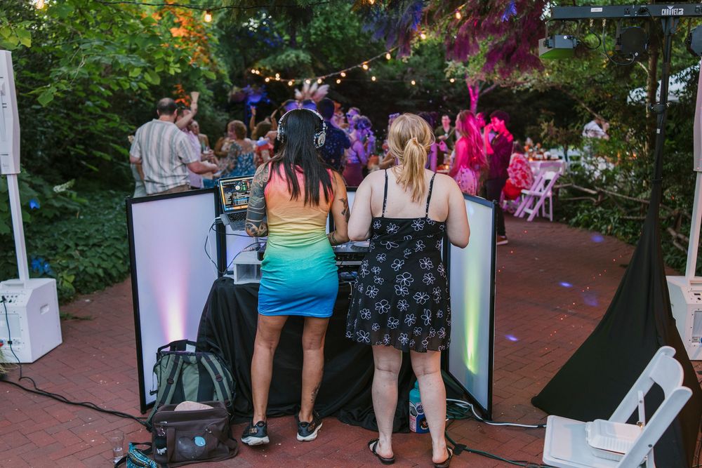 A group of party guests gather around a DJ booth at an outdoor garden event with string lights overhead.
