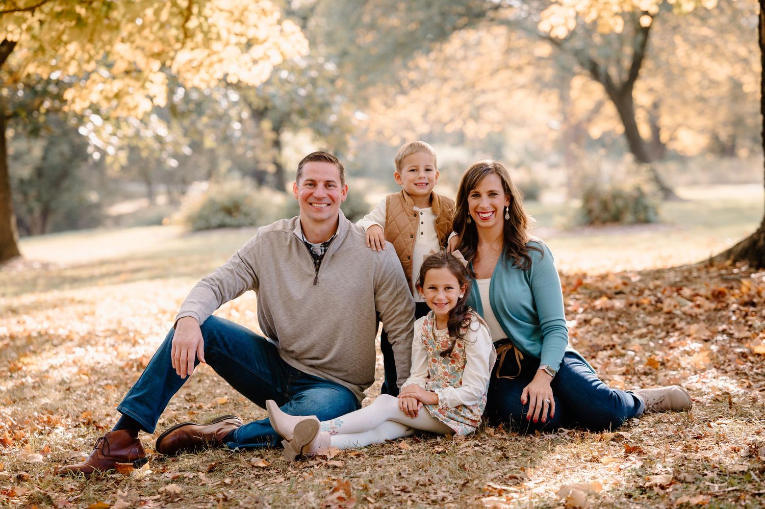 A family poses together on autumn leaves in a sunlit park during golden hour.