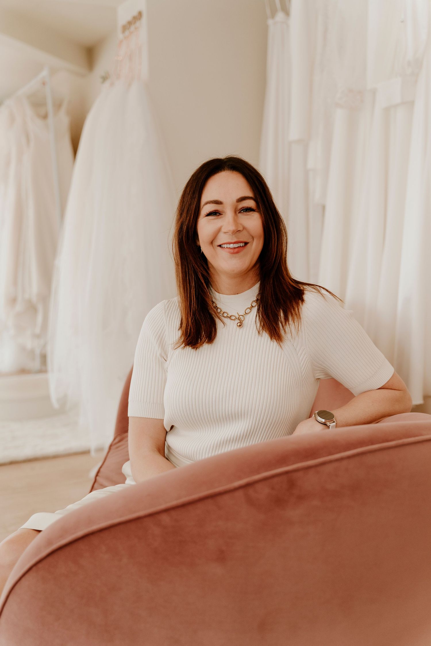 Someone in a white ribbed sweater sits on a pink couch in a bridal boutique with wedding dresses visible in background.