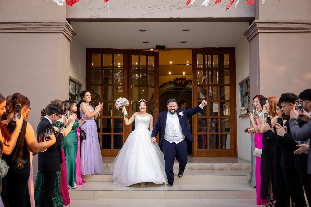 Wedding party exits church entrance while guests in colorful attire cheer and take photos.