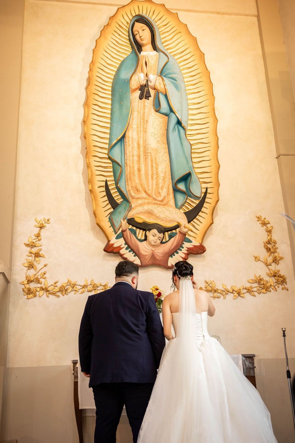 A couple prays before a large Virgin of Guadalupe mural in a church.
