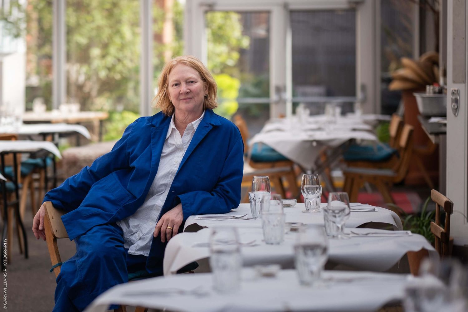 Someone in a bright blue blazer sits comfortably at an outdoor cafe table set with white linens and glassware.