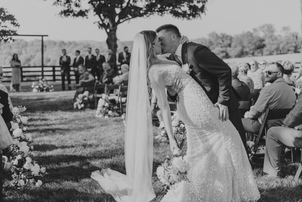 Black and white photo of a romantic outdoor wedding ceremony kiss with guests seated on lawn and trees in background.