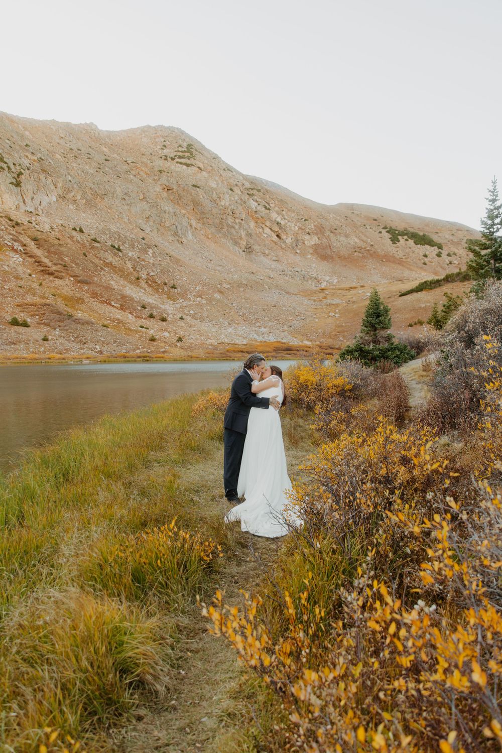 A couple embraces by a serene mountain lake surrounded by autumn foliage and rugged terrain on their wedding day in Denver, Colorado.