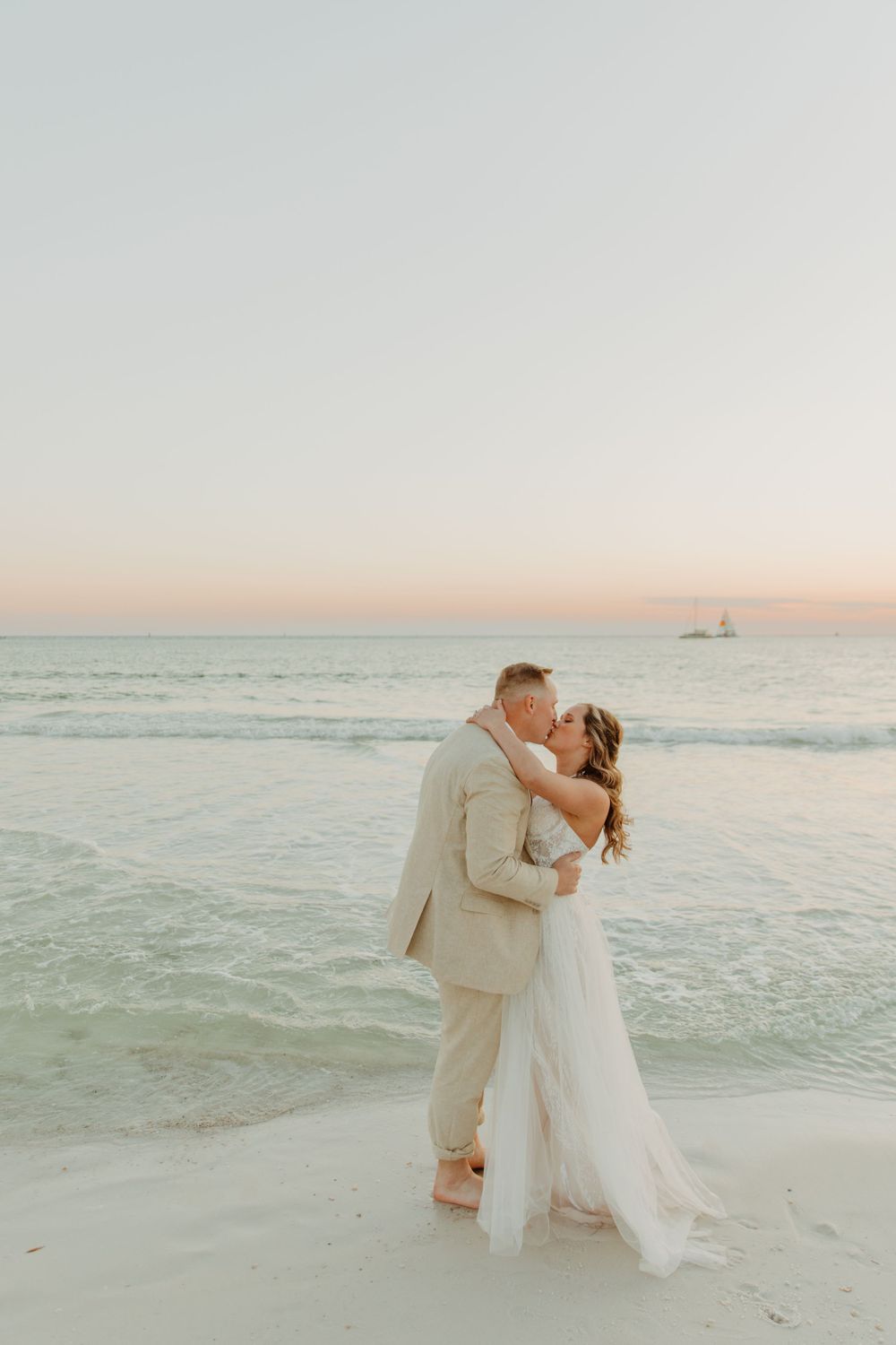 A couple shares a romantic kiss on a beach at sunset with soft pastel colors in the sky and ocean waves behind them in Panama City Beach, FL