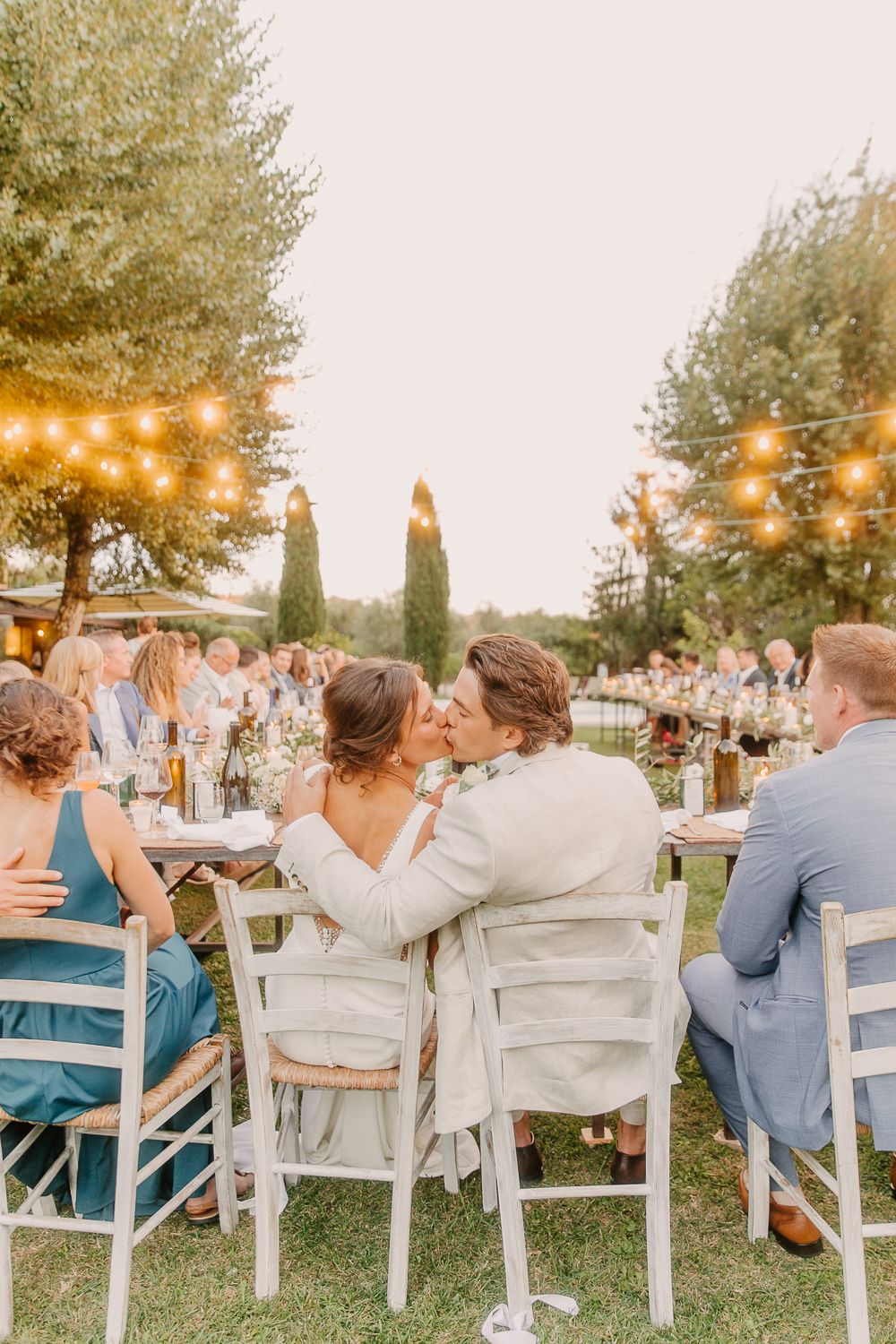 Guests seated at outdoor wedding reception with string lights overhead and greenery in background at sunset in Tuscany, Italy.